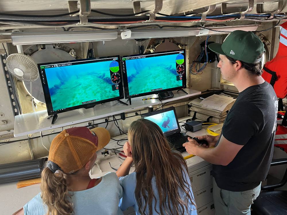 an adult and two children in a ship control room look at screens with a display showing live underwater video