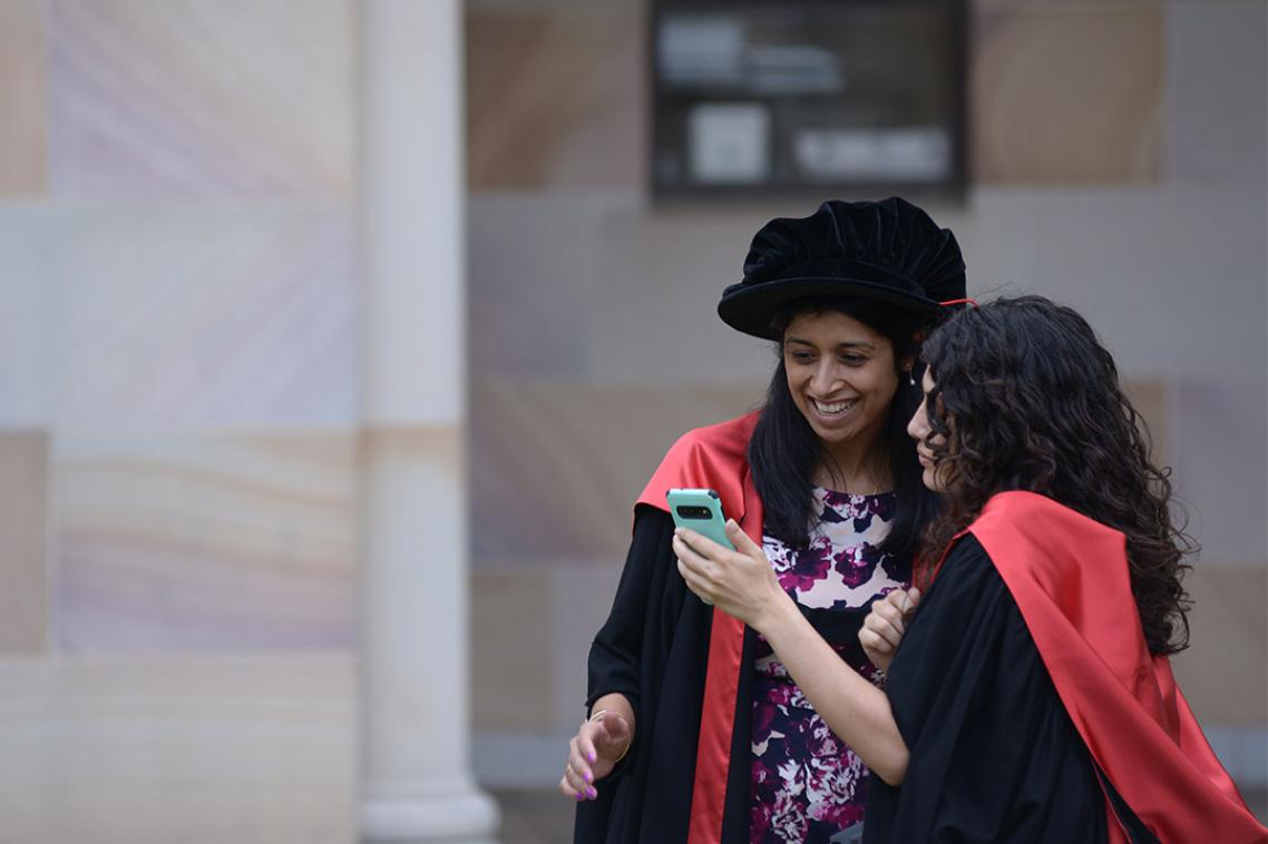 Doctoral graduate in gown and graduation tam, looking at phone with friend