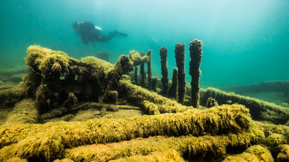 a diver floats above a shipwreck in the distance with debris from the wreck in the foreground