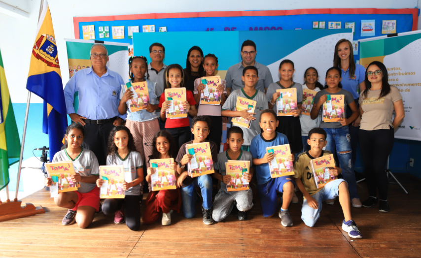 Várias crianças seguram livros didáticos em uma sala de aula. Ao lado estão adultos.