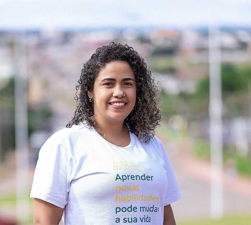 Uma mulher de cabelo encaracolado sorri para foto. Ela veste uma camiseta com a frase “Aprender novas habilidades pode mudar a sua vida”