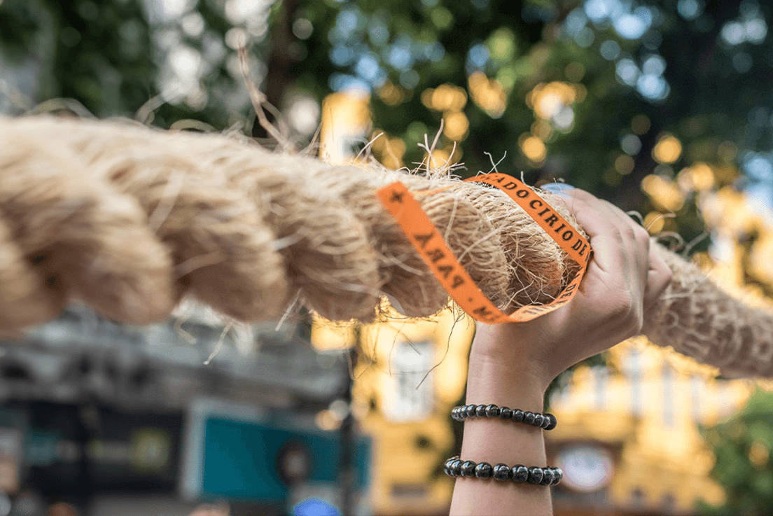 Imagem de uma mão segurando a corda do Círio. Na corda, há uma fitinha laranja 