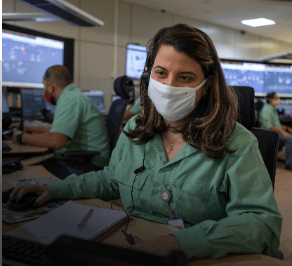 Photo of a woman sitting on a chair in an office with several monitors and screens in the background. She has dark shoulder-length hair with highlights and is wearing a uniform, a green button-up blouse, and a face mask.