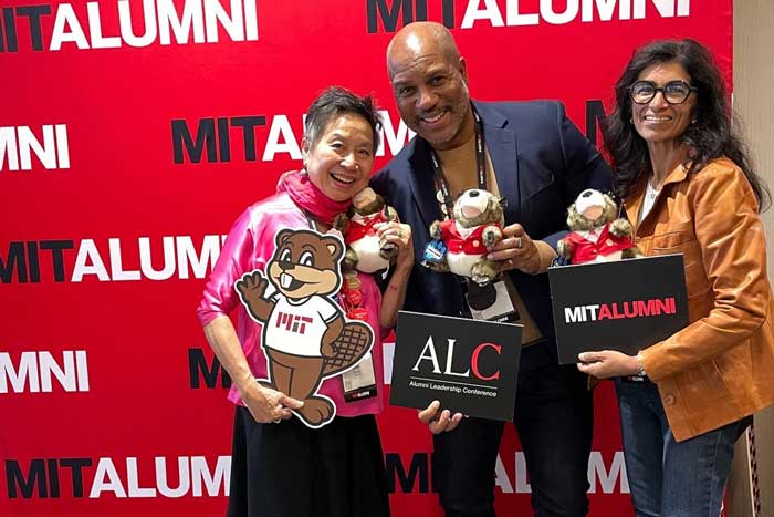 3 people pose with MIT merch and Tim plushies in front of MIT Alumni banner.