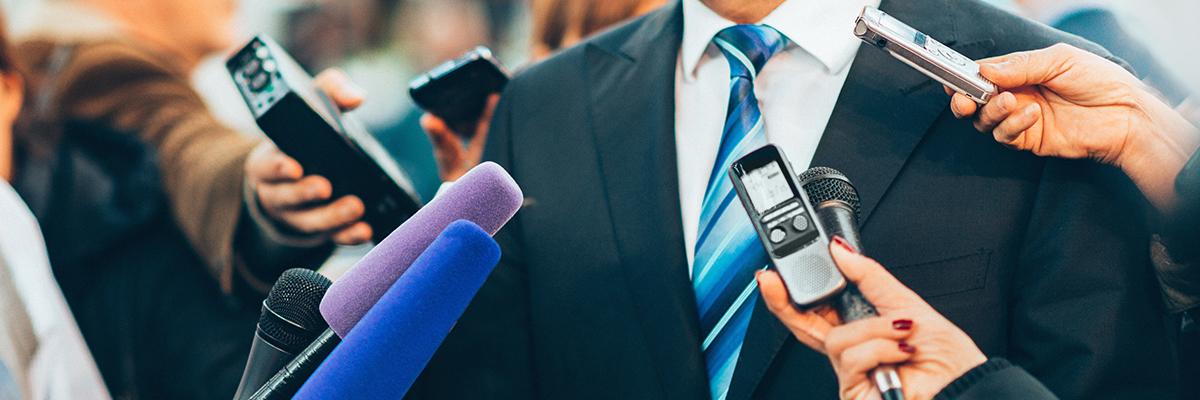 faceless figure in suit and tie surrounded by reporters' microphones and recording devices