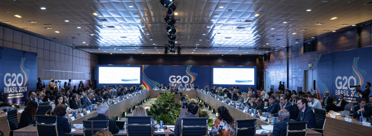 Delegates seated around a large table at the G20 Brasil 2023 conference room, with banners and screens displaying the event's name.