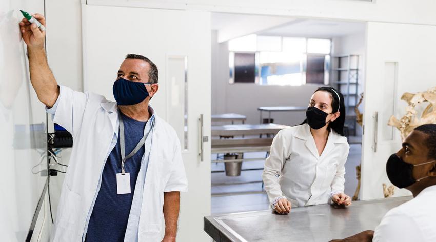 A masked professor teaches two masked students in a classroom