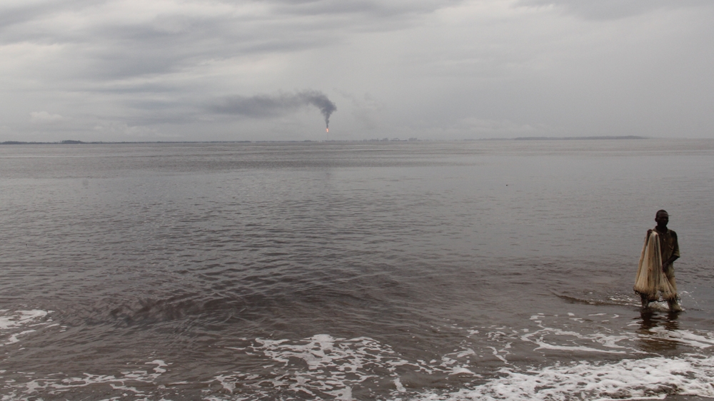 A man fishes on a beach at Banana Point as smoke is emitted from Angola's massive oil industry in the distance [William Clowes/Al Jazeera]