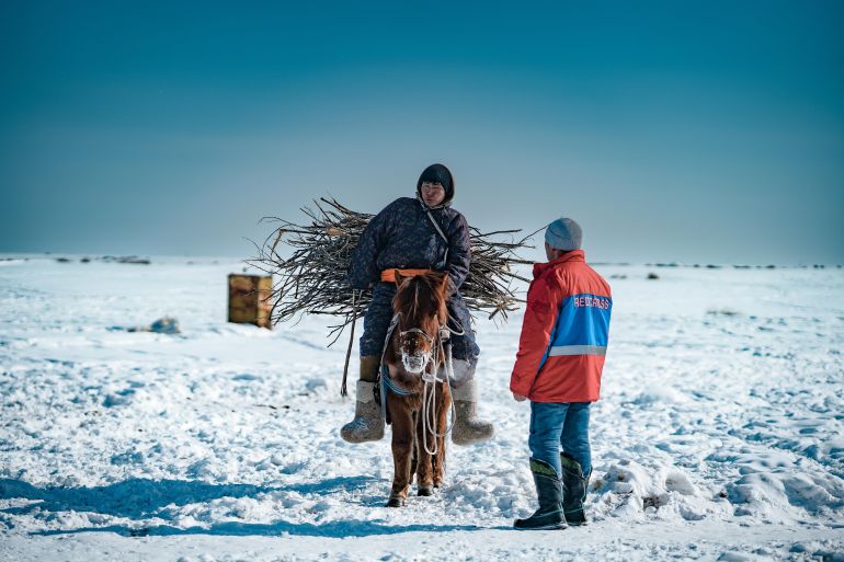 a man on a donkey with sticks talks to another man in the snow