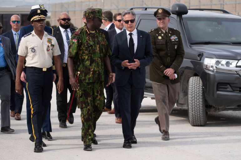 Antony Blinken — in a suit and sunglasses — walks alongside men in military fatigues at a Port-au-Prince base.