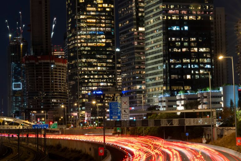 Traffic moves along a road in front of tall buildings at night