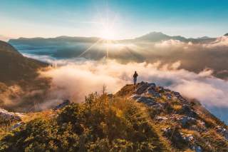 Person auf Bergkuppe vor Bergkulisse mit Sonnenschein und Wolken im Tal