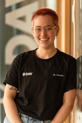 Person mit kurz rasierten pinken Haaren und einer Brille. Die Person trägt ein schwarzes T-Shirt mit dem DAV Logo und eine hellblaue Jeans.