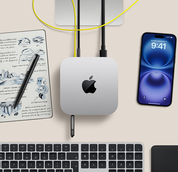 Top view of silver Mac mini with cords and a flash drive plugged into ports, in a desk setup with wireless keyboard, trackpad, iPhone, and notepad