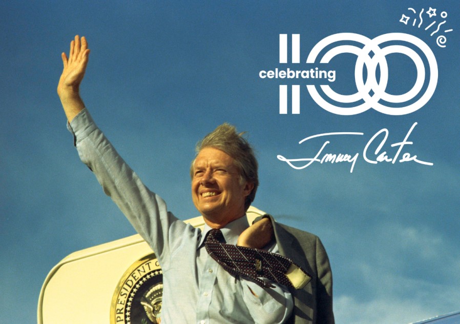 Color photograph of President Carter waving in front of a plane with the Celebrating 100 Jimmy Carter logo in the upper right corner