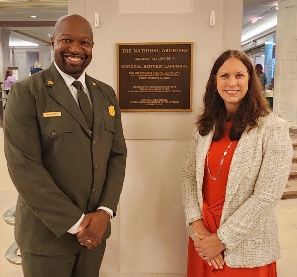 Color photograph of a Black man in a National Park Service uniform and a White woman in a red dress standing on either side of a plaque