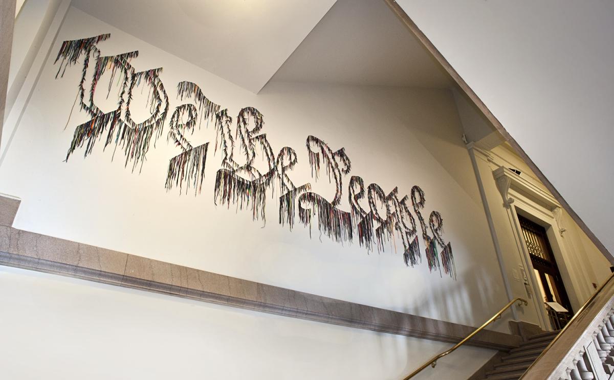 color photograph of art installation on a white wall that spells "We the People" in shoelaces