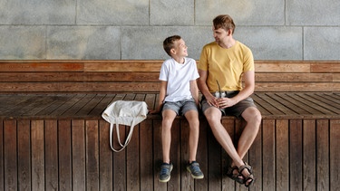 Vater und Sohn unterhalten sich, beide sitzen auf einer Holzbank. Was bedeutet Gewaltfreie Kommunikation mit Kindern? Der Psychologe Marshall B. Rosenberg hat sie entwickelt. Wir erklären, warum Gewaltfreie Kommunikation mit Kindern wichtig ist, wie ihr sie in vier Schritten erlernen könnt und was es mit der Giraffensprache auf sich hat.  | Bild: picture alliance/Westend61/Ekaterina Yakunina