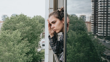 Eine junge Frau lehnt sich besorgt aus dem Fenster eines Hochhauses | Bild: colourbox.com