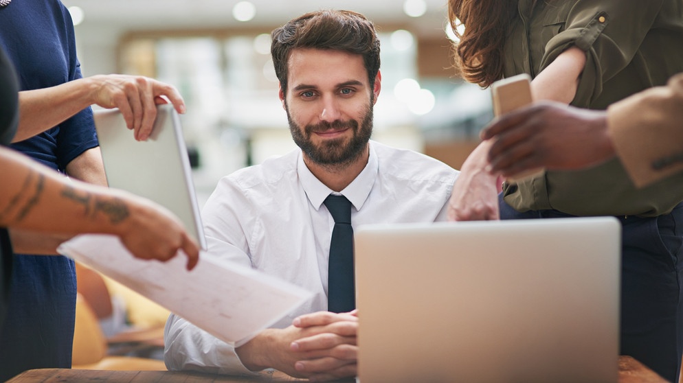 Mann sitzt an Schreibtisch, mehrere Hände reichen ihm Hefte. Multitasking kann zum Burnout führen.  | Bild: colourbox.com