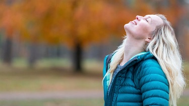 Eine Frau steht im Park und lächelt mit geschlossenen Augen. Selbstliebe bedeutet, mit sich selbst befreundet zu sein, zu seinen Bedürfnissen und Wünschen zu stehen und die eigenen Schwächen zu akzeptieren. Wie du lernst, dich selbst zu lieben. | Bild: picture alliance / Westend61 | William Perugini