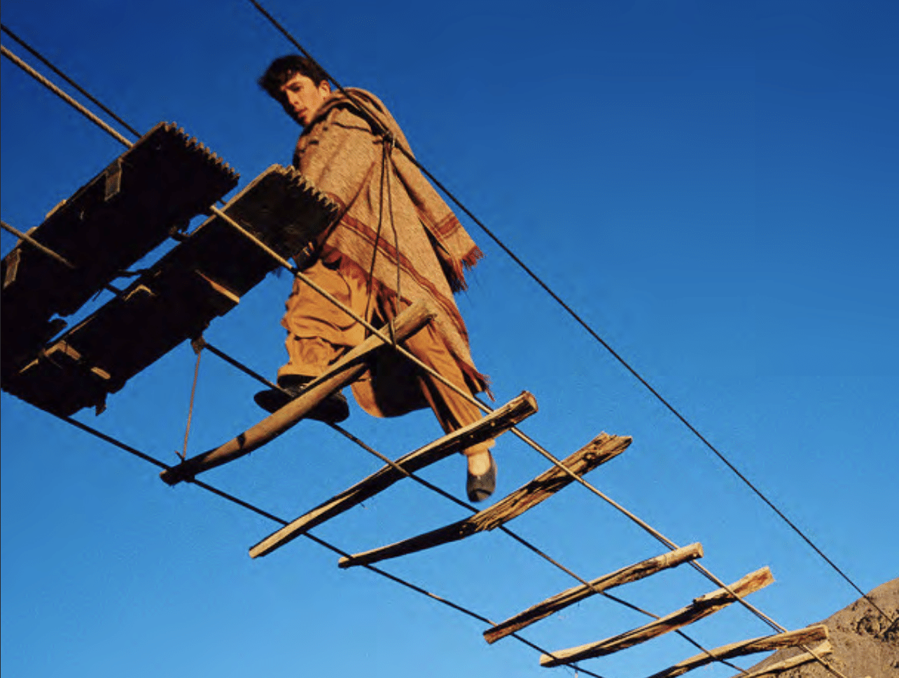 Person in a cloak walking across a bridge that has minimal wooden boards on it