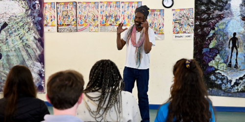 Students listening to someone giving a talk in front of a wall of artwork.	