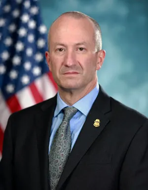 Troy A. Miller, CBP Senior Official Performing the Duties of the Commissioner positioned in front of a blue background with a flag to the left.