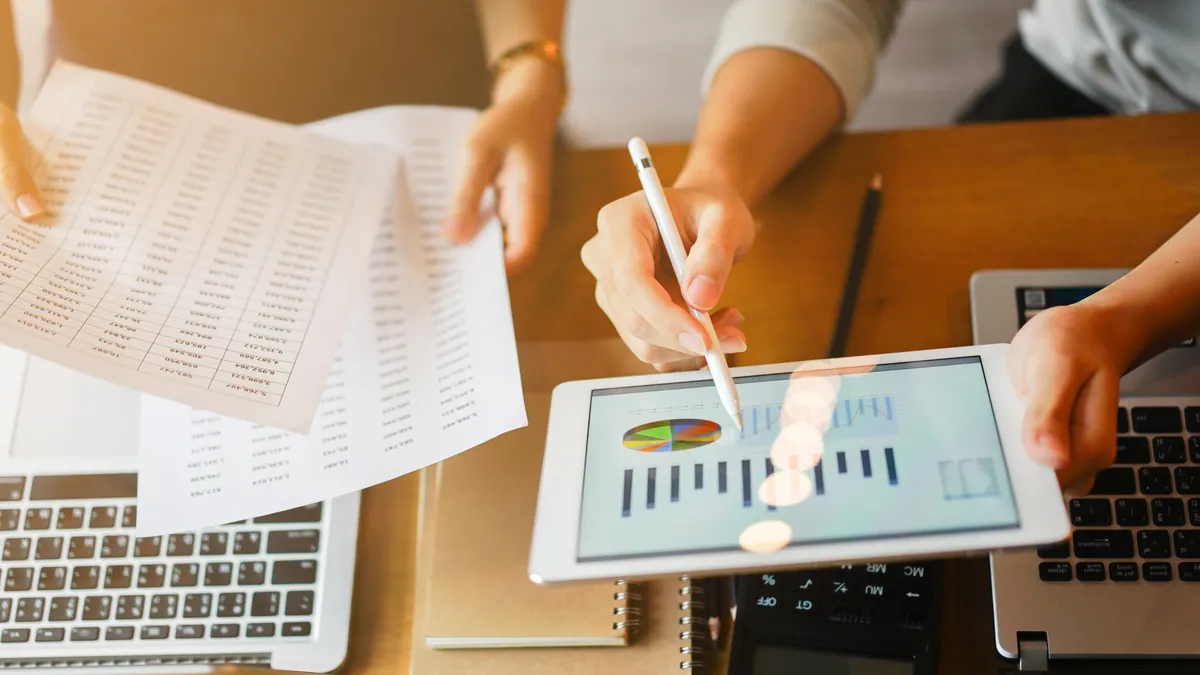 Businessman holds electronic tablet showing profit data, while businesswoman holds hard-copy report.