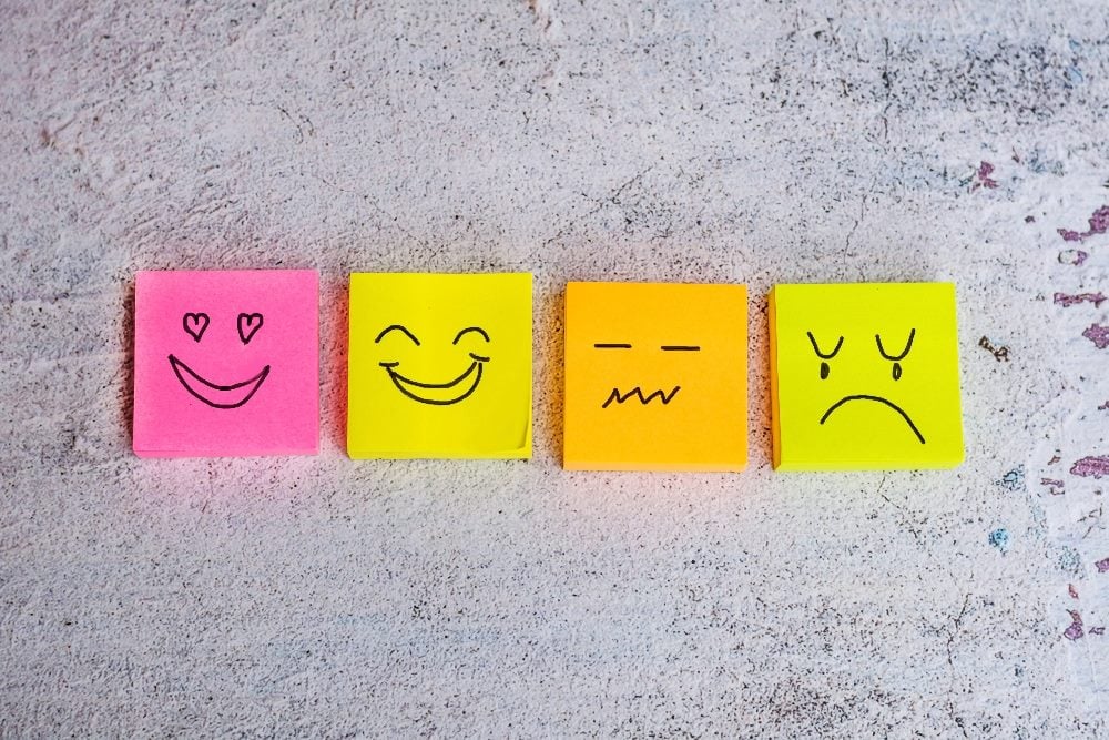 Hand-drawn faces of happy, sad and neutral emotions on colorful, pink, yellow, orange and green, sticky notes, lying on a mottled surface in piece about customer support in CX.