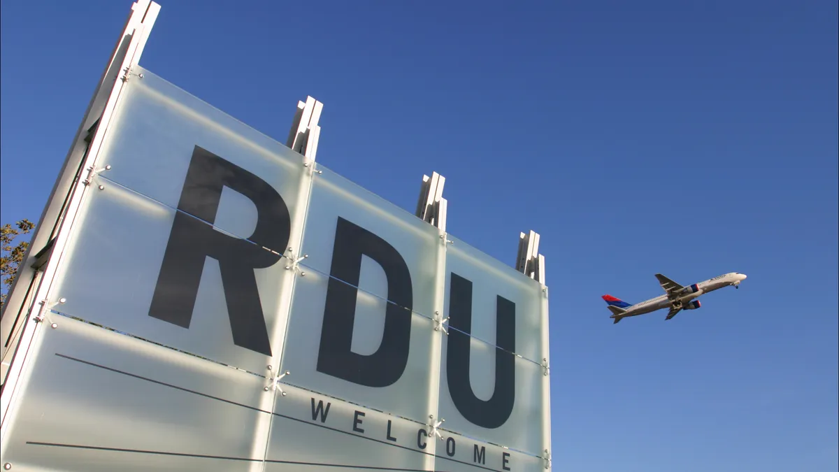 A photo of the Raleigh-Durham International Airport in North Carolina. A plane is flying in the background. In front is a grey building with "RDU" on the front in black letters.