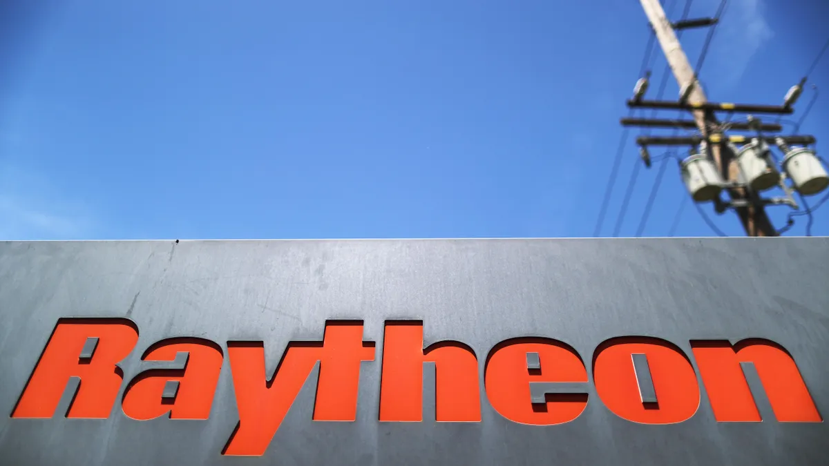 With a blue sky in the background, a black sign that reads "Raytheon" in large red letters.