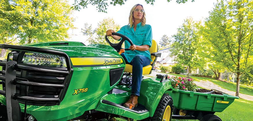 Women on lawn mower hauling flowers and mulch with her cart attachment