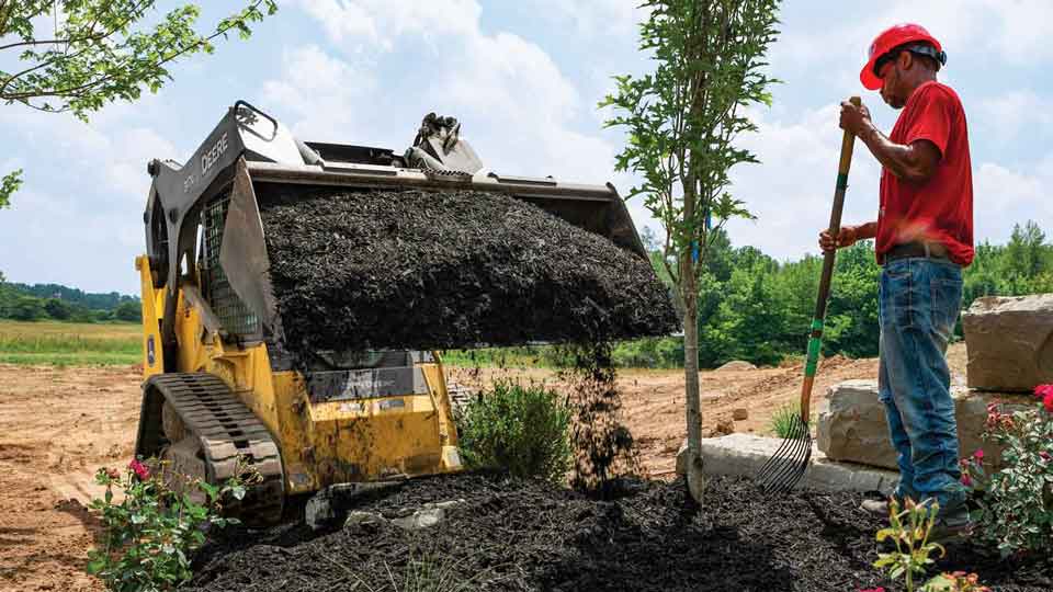 John Deere 317G Compact Track Loader with bucket attachment dumping dirt into a garden with a man with a man and a shovel standing by