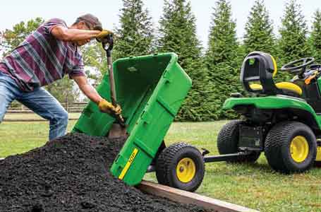 Man scooping soil out of a 10P poly cart that is attached to a John Deere Lawn Tractor