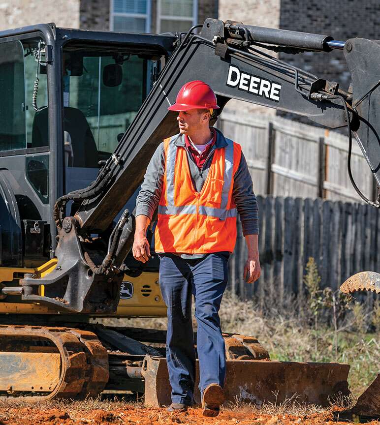 a man in a hard hat and vest walking in front of John Deere construction equipment on a job site.