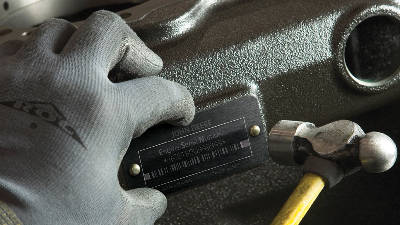 A person wearing a gray glove holding a hammer while installing a John Deere engine serial number plate on an engine