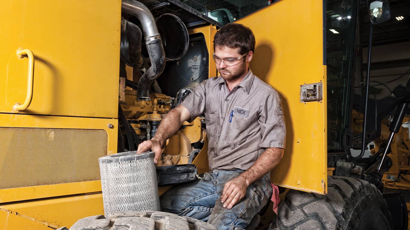 A man wearing safety goggles working on a John Deere engine inside of a heavy equipment machine