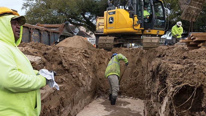 Four employees with their backs to the camera watch a John Deere 380G LC Excavator, a 460E Articulated Dump Truck, and an 872 GP SmartGrade Motor Grader work at moving dirt.