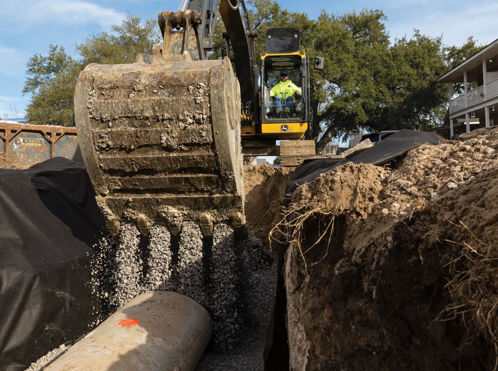 An excavator drops gravel on a buried pipe laying in a trench