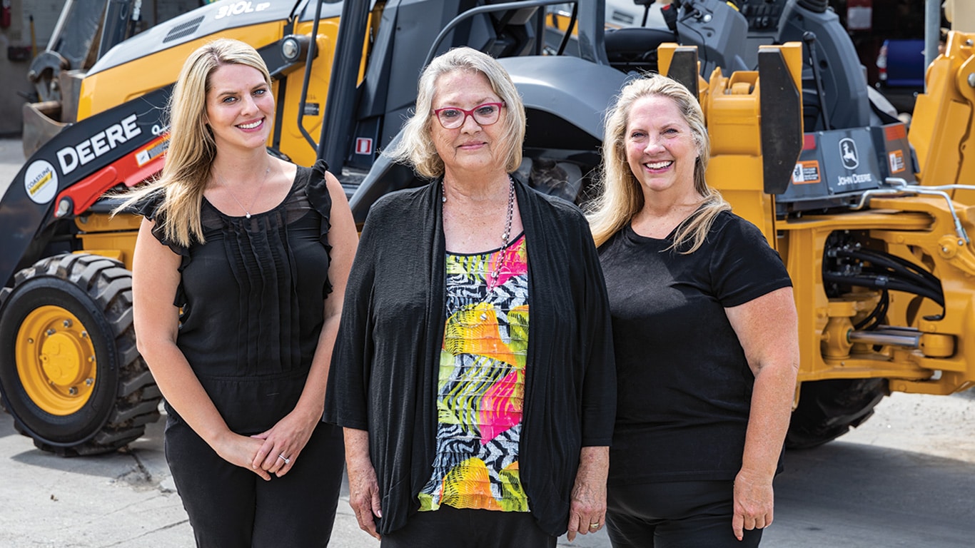 Representing three generations, Amanda Matson, Janet Ferguson, and Nikki Worden of Newman Backhoe Service stand in front of a John Deere 310L Backhoe.