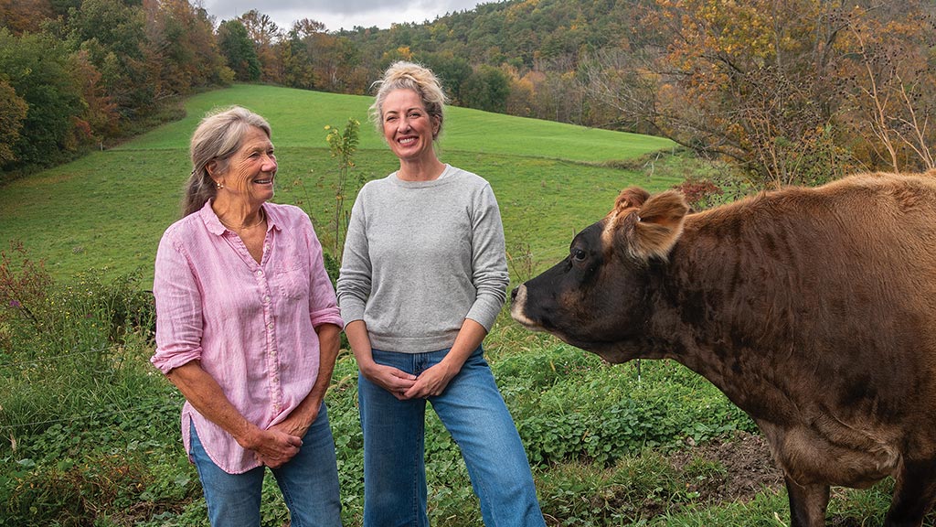 Two smiling people standing next to a brown cow with a green hillside rising behind them 