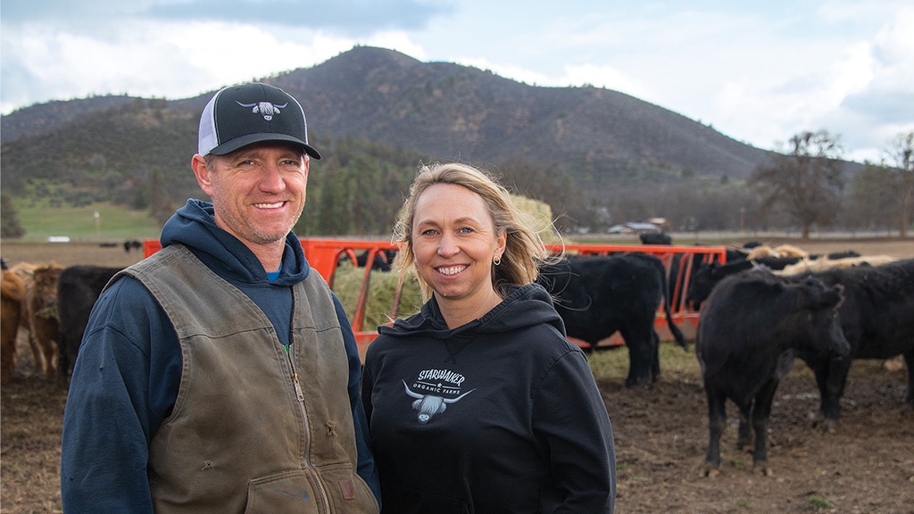 Two smiling people standing in front of feeding black and brown cows with a hill in the distance 