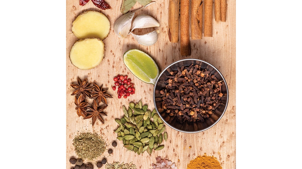 A cutting board with cloves, star anise, sliced ginger, garlic cloves, green cardamom, red peppercorns, fennel seed, curry powder, lime, pink salt, cinnamon sticks, bay leaf, and dried red chilies on it