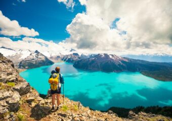 Backpacker stands on a cliff overlooking a blue lake and a mountains in the distance.