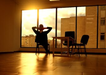 Bueiness professional with their hands behind their head looking out office window towards sun above nearby buildings.