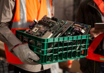 Working in orange safety vest carrying a green bin of sorted e-waste components for recovery and recycling.