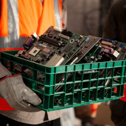 Working in orange safety vest carrying a green bin of sorted e-waste components for recovery and recycling.