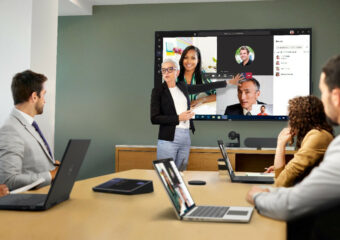 Woman leads presentation in office meeting room in front of large display showing colleagues who are taking part in the meeting from remote locations.