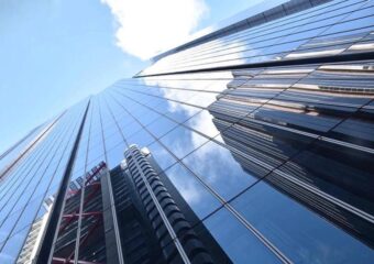 Reflections of high rise buildings appearing in the glass windows of another high rise building with a partly cloudy sky above.